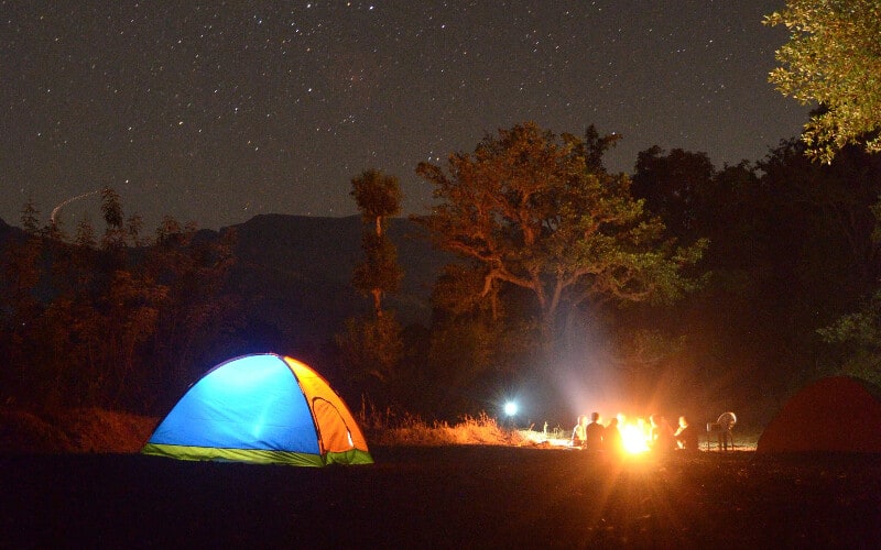 A camping tent lit up next to a group of people around a campfire at night.