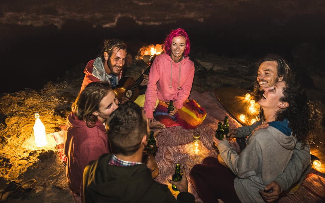 A group of friends having fun on the beach using campsite lighting ideas such as lanterns, fire, and bottles with lights to illuminate their surroundings.