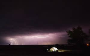 A tent lit up at night in the middle of the field with lightning in the distance.