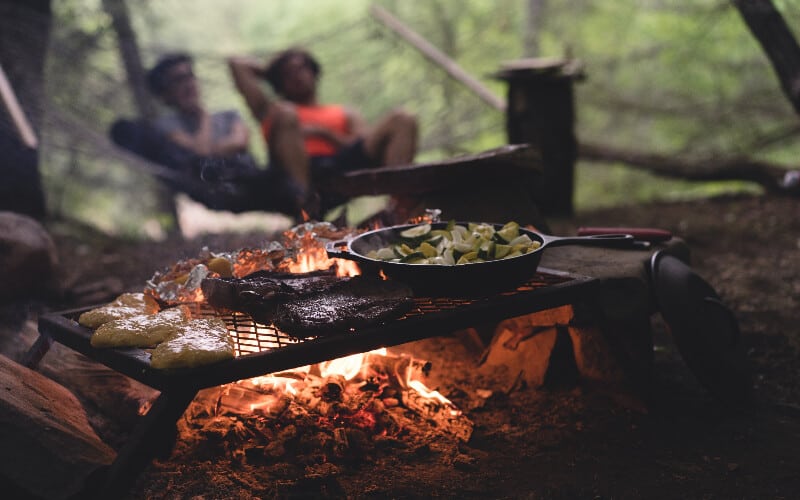 Food cooking on a grill over a campfire with two people in a hammock in the background.