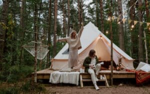 A man playing a ukulele while a woman dances in front of their tent on a glamping trip. 