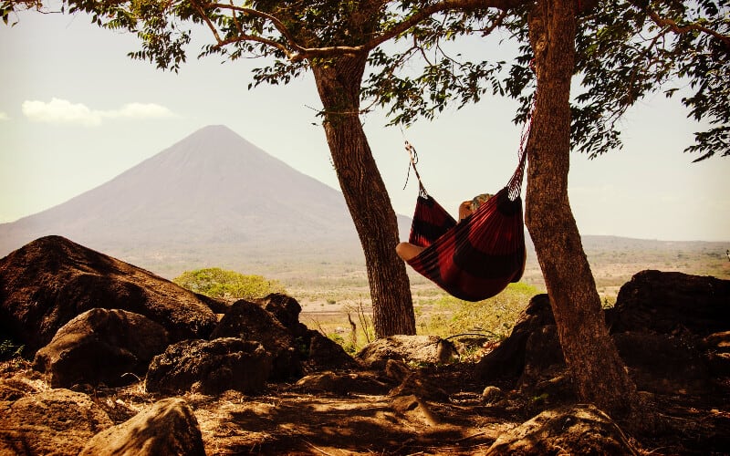 A person relaxing in a hammock between two large trees with a mountain in the distance.