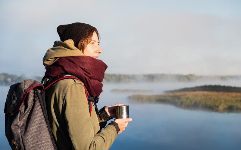 A woman staying warm with her hiking scarf while sipping a warm drink and overlooking a foggy river.