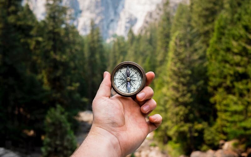 A hand holding a compass on a hike with trees in the background.